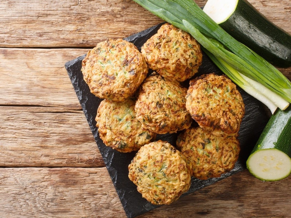 Eine dunkle Platte mit Kolokithokeftedes neben frischer Zucchini darauf.