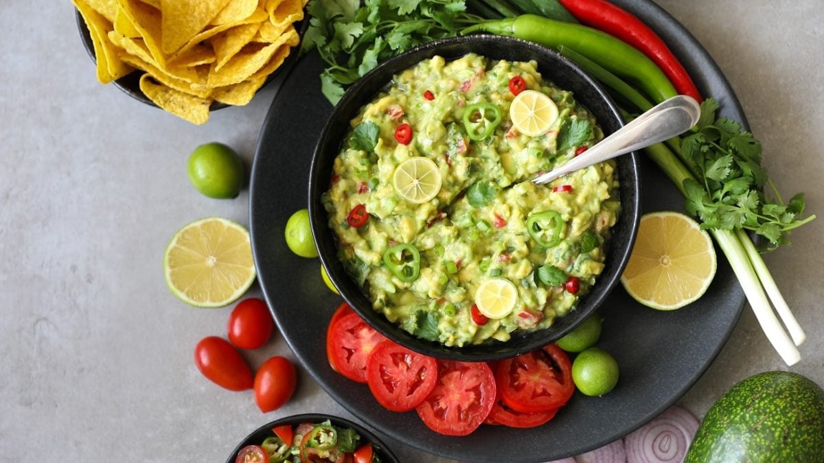 Draufsicht: eine dunkle Schüssel auf hellem Grund. Darin ist eine Avocado-Koriander-Salsa mit frischen Chiliringen. Um den Teller herum liegen frischie Chili, Tomatenscheiben und Nachos.