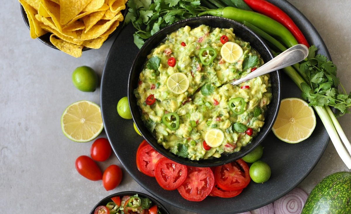 Draufsicht: eine dunkle Schüssel auf hellem Grund. Darin ist eine Avocado-Koriander-Salsa mit frischen Chiliringen. Um den Teller herum liegen frischie Chili, Tomatenscheiben und Nachos.