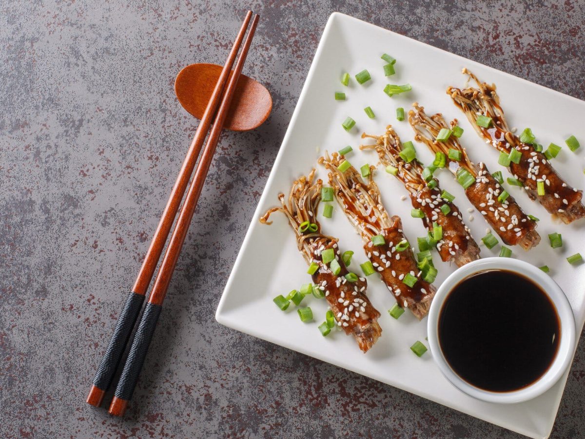 Ein weißer quadratischer Teller mit eine rkleinen Schüssel mit dunkler Sauce und den beef-Enoki-Rolls auf einem grauen Tisch.