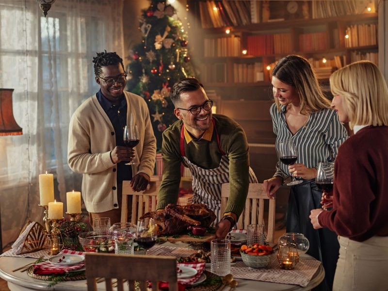 Festliche Szene mit vier Menschen, die um einen reich gedeckten Tisch voller Speisen stehen, Weihnachtsbaum und Lichter im Hintergrund.