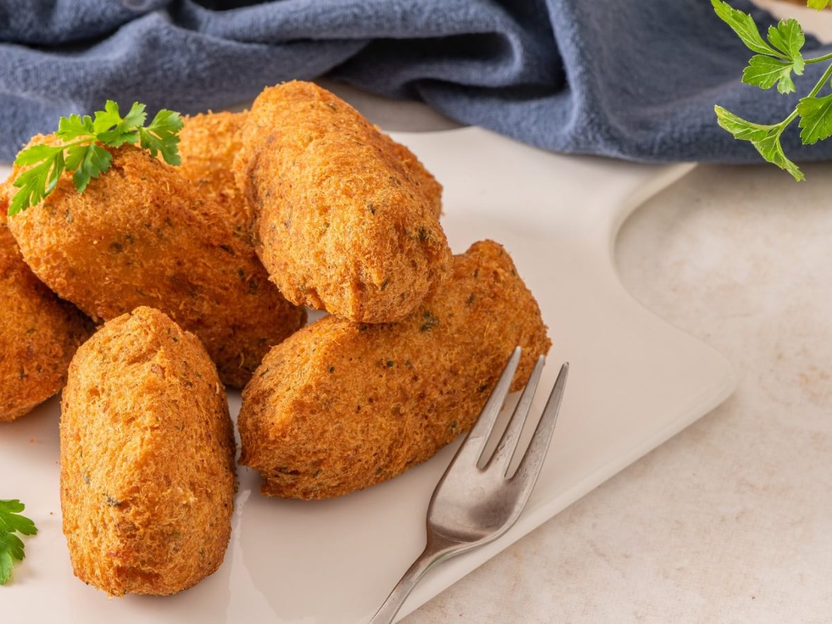 Einige Fischkroketten auf einem weißen Teller mit einem blauen Tuch im Hintergrund. Daneben noch ein Zweig Petersilie.