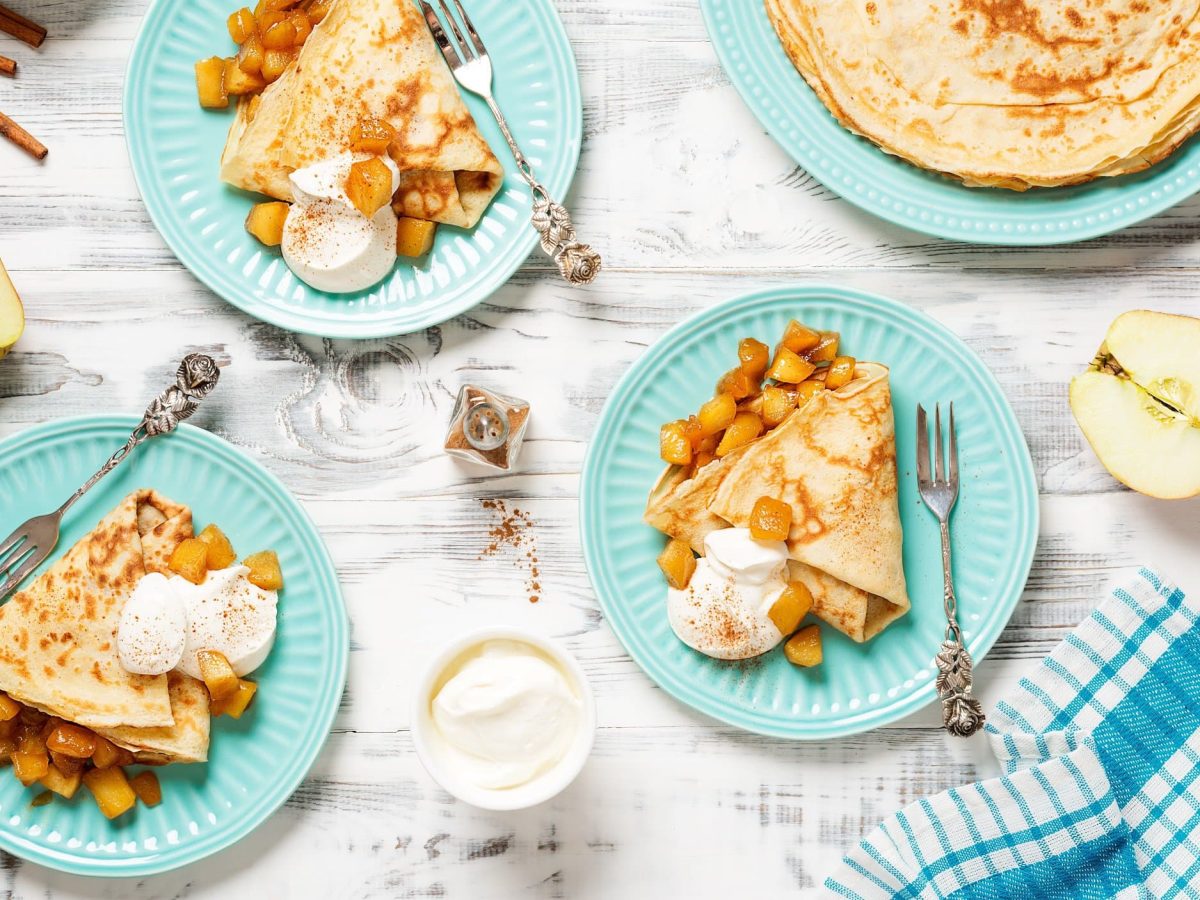 Drei Teller mit Bratapfel-Eierkuchen und Sahne in der Draufsicht, daneben ein großer Teller mit weiteren Eierkuchen, halbierte Äpfel und Zimtstangen.