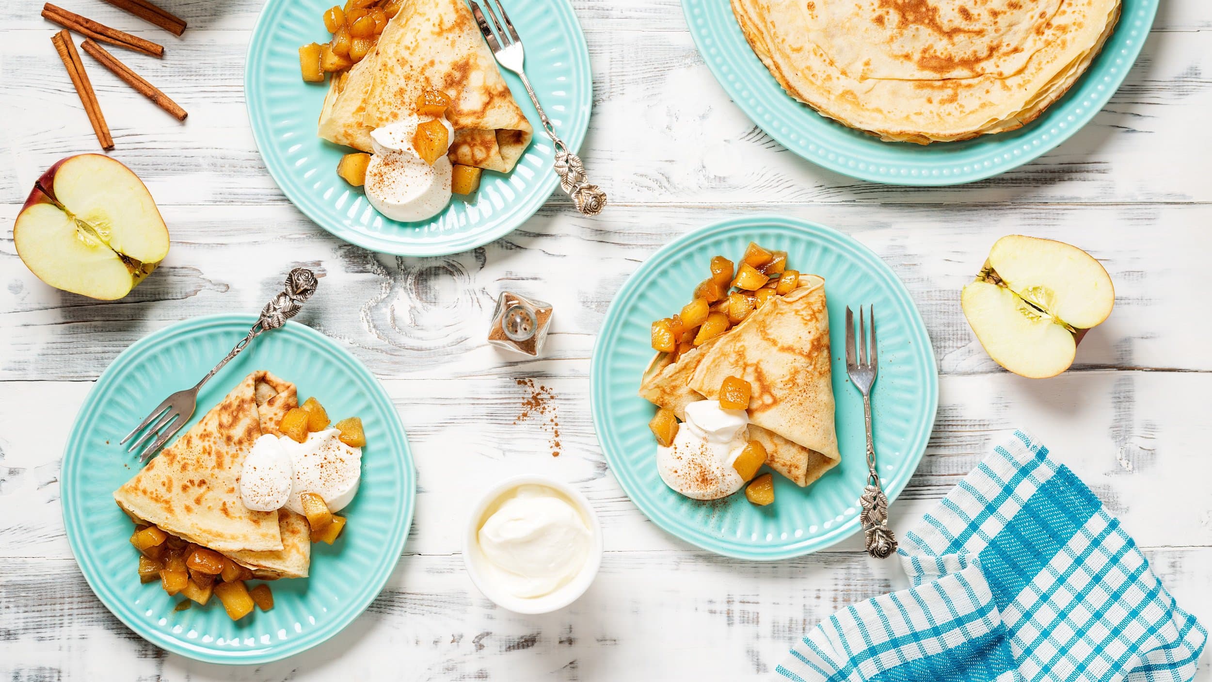 Drei Teller mit Bratapfel-Eierkuchen und Sahne in der Draufsicht, daneben ein großer Teller mit weiteren Eierkuchen, halbierte Äpfel und Zimtstangen.