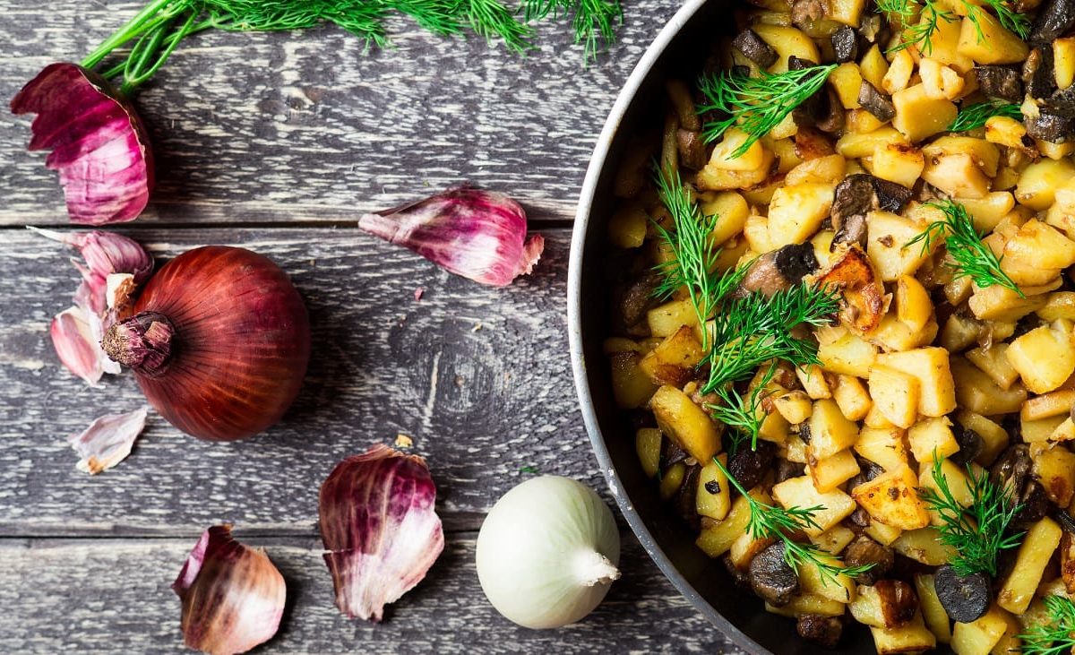 Draufsicht: Auf einer groben Holzplatte liegen eine rote Zwiebel, Zwiebelschalen sowie eine weiße Zwiebel. Daneben steht eine Pfanne mit Bratkartoffeln und Birne.