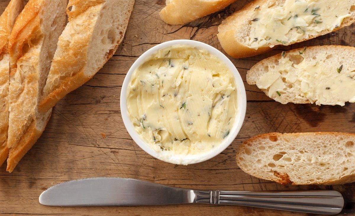 Draufsicht: eine helle kleine runde Schale mit einer kräuterbutter auf einem Untergrund auf Holz. Drum herum lieben aufgeschnittene baguettescheiben.