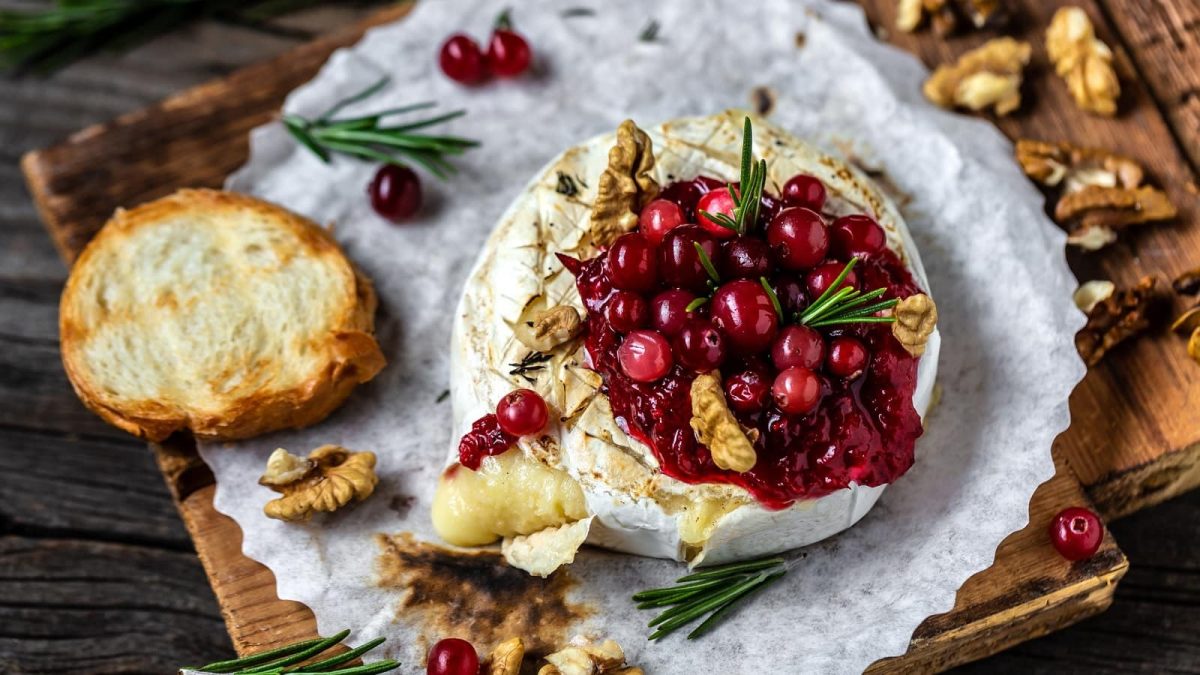 Ein runder gebackener Camembert mit einer roten Crannberysauce und gerösteten Walnüssen. Liegend auf einem Stück Backpapier und Holz. Serviert mit einer Scheibe geröstetem Brot.