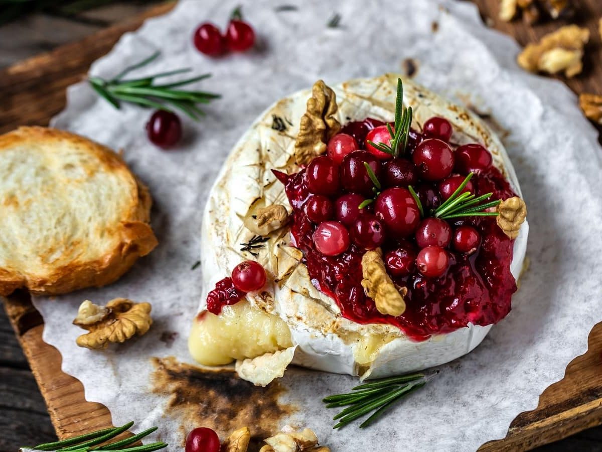 Ein runder gebackener Camembert mit einer roten Crannberysauce und gerösteten Walnüssen. Liegend auf einem Stück Backpapier und Holz. Serviert mit einer Scheibe geröstetem Brot.