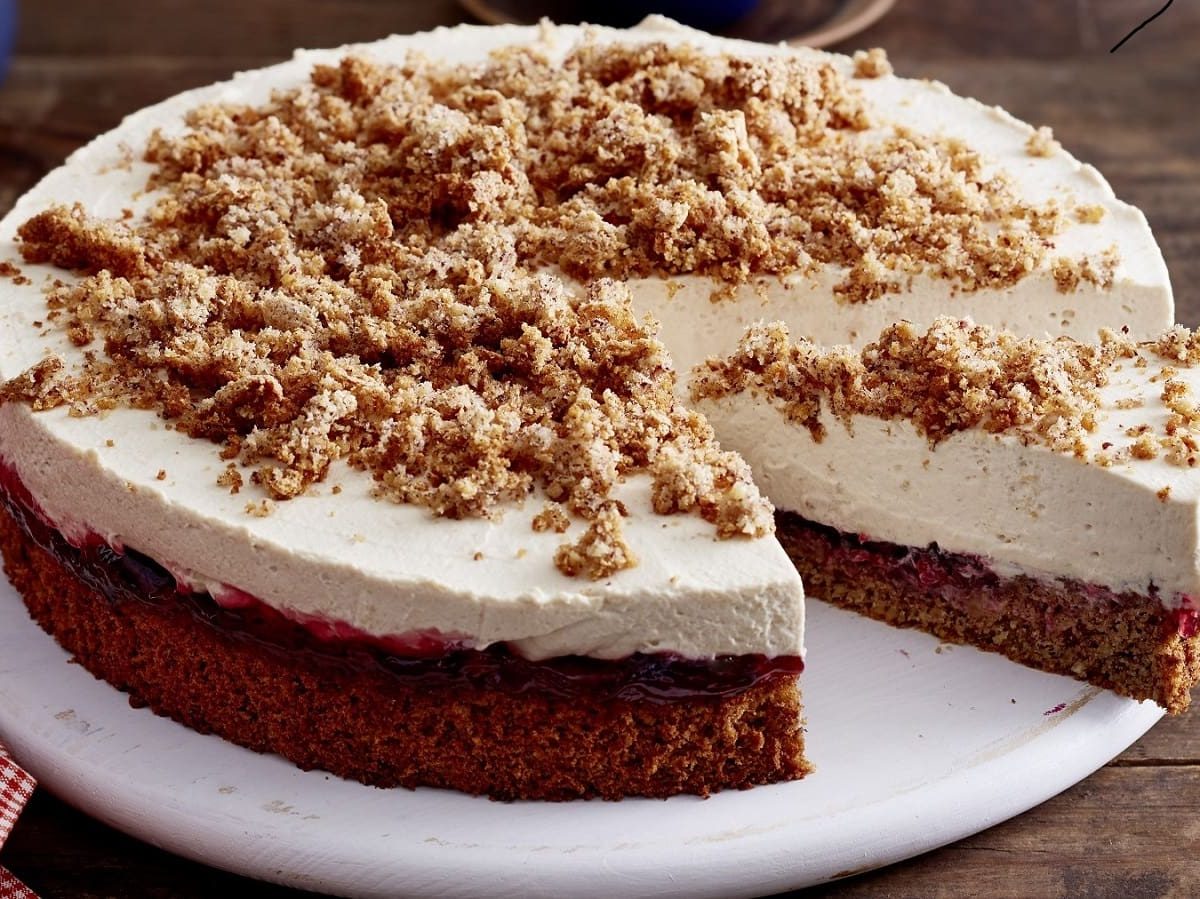 Angeschnittene Torte auf einem weißen flachen Teller auf einem hölzernen Untergrund. Die Torte hat einen braunen Boden und eine helle Cremeschicht, die mit Krümeln dekoriert ist.