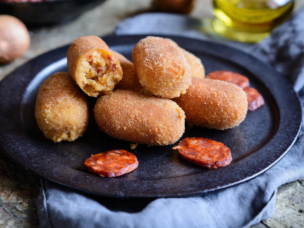 Ein dunkler Teller mit den Chorizo-Kroketten auf einem grauen Marmoruntergrund mit blauem Tuch.