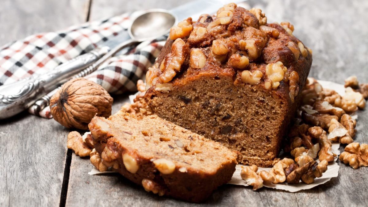 Ein angeschnittenes Dattel-Nuss-Brot auf einem hellen Holztisch mit eine rrot-schwarz-weiß-karierten Decke.