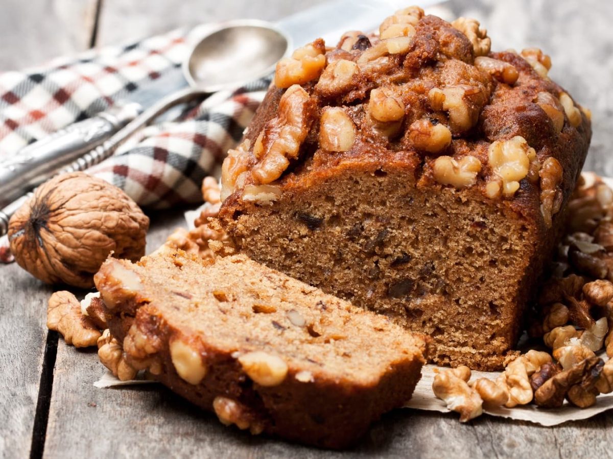 Ein angeschnittenes Dattel-Nuss-Brot auf einem hellen Holztisch mit eine rrot-schwarz-weiß-karierten Decke.