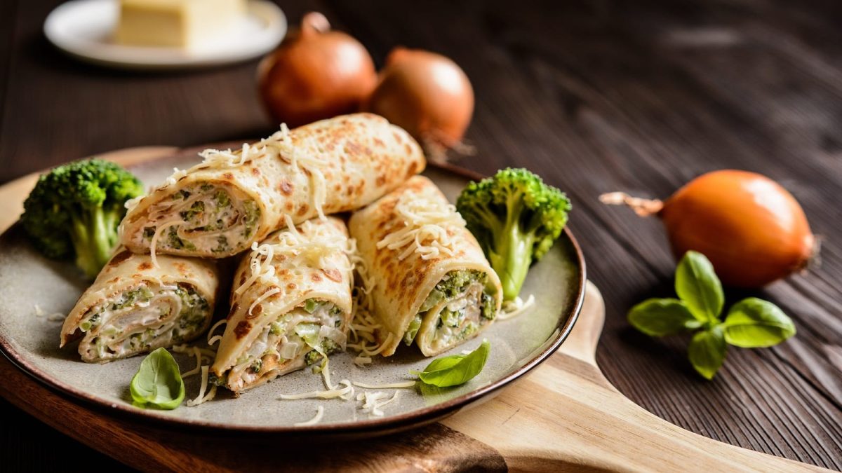 Ein Teller mit gefüllten Crêpes-Röllchen mit Brokkoli auf einem Holzbrett.