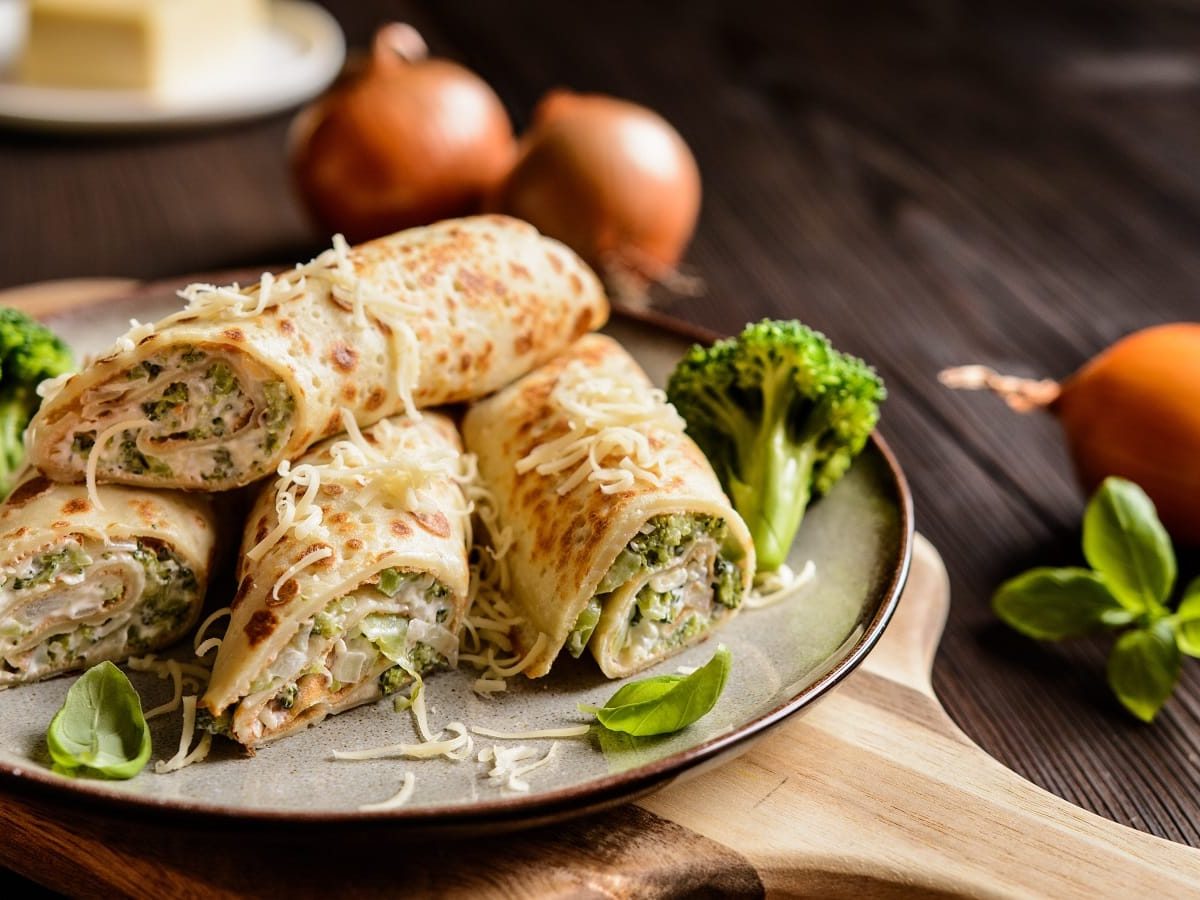 Ein Teller mit gefüllten Crêpes-Röllchen mit Brokkoli auf einem Holzbrett.