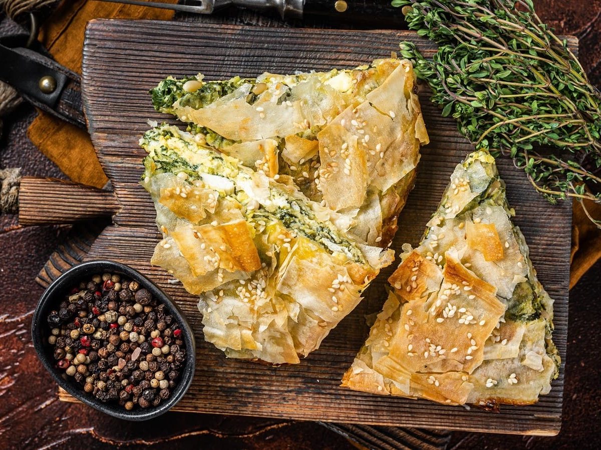 Drei Stücke Spanakopita auf einem Holzbrett neben einer Schale mit Pfefferkörnern.