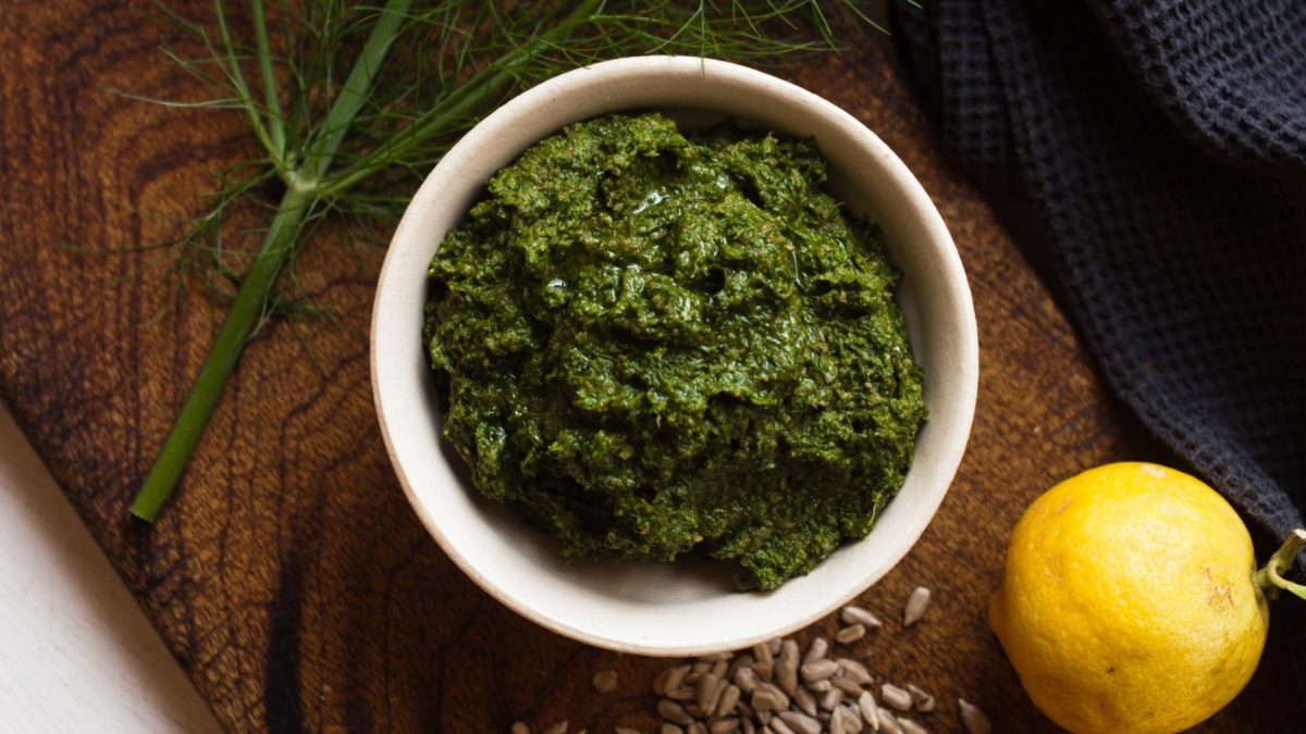 Fenchel-Pesto in einer kleinen Schale angerichtet auf einem Holzbrett, dazu serviert frisches Fenchelgrün und Zitrone.