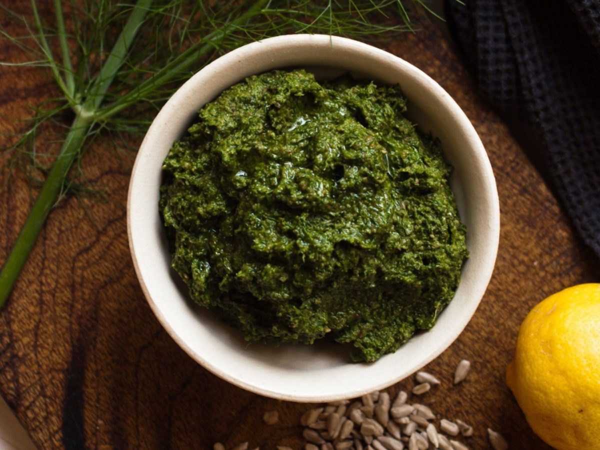 Fenchel-Pesto in einer kleinen Schale angerichtet auf einem Holzbrett, dazu serviert frisches Fenchelgrün und Zitrone.