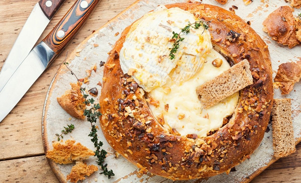 Draufsicht: Ein rundesbrot ist in der Mitte ausgehölt. In dem Loch ist ein zerlassener Camembert-Käse. Auf ihn ist ein Zweig Thymian. Drum herum liegen brotstücke und zwei Messer mit Schäften aus Holz.