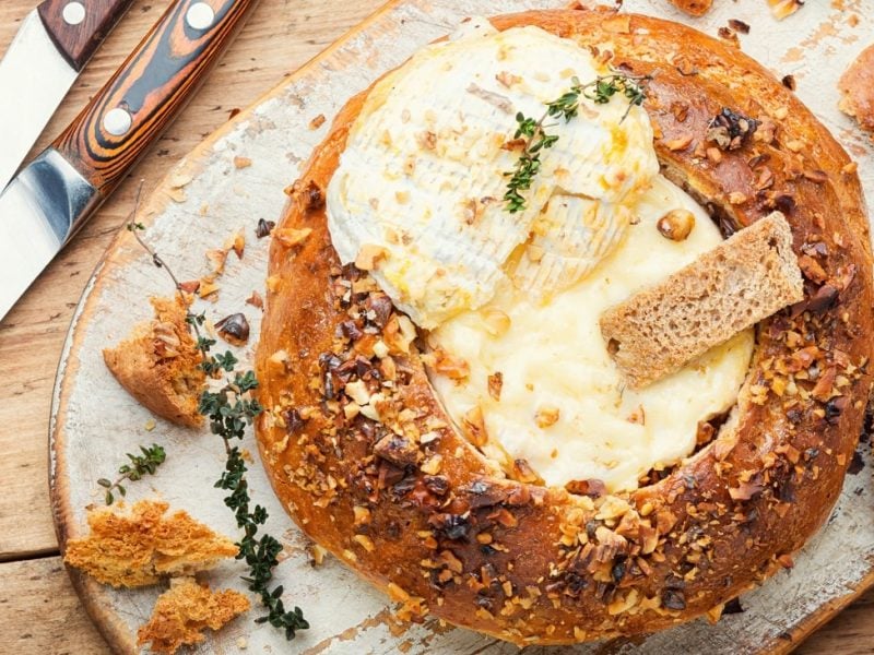 Draufsicht: Ein rundesbrot ist in der Mitte ausgehölt. In dem Loch ist ein zerlassener Camembert-Käse. Auf ihn ist ein Zweig Thymian. Drum herum liegen brotstücke und zwei Messer mit Schäften aus Holz.