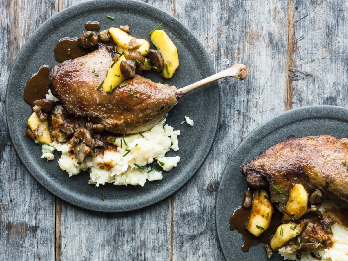 Gänsekeulen mit Äpfeln und Maronen auf Reis, serviert auf Keramiktellern auf einem grauen Holztisch.