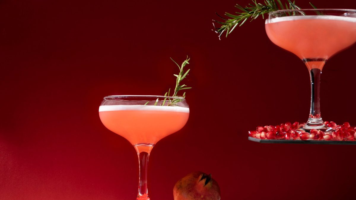 Zwei Granatapfel Vodka Sour Flamed Rosemary in Cocktailschalen vor rot-schwarzem Hintergrund. Einer erhöht auf Etagere. Im Bild Granatapfelkerne. Frontalaufnahme.