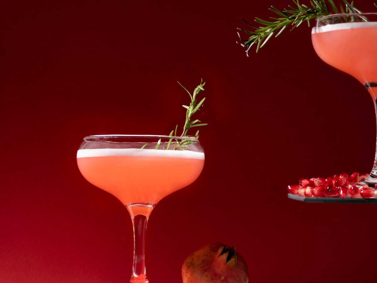 Zwei Granatapfel Vodka Sour Flamed Rosemary in Cocktailschalen vor rot-schwarzem Hintergrund. Einer erhöht auf Etagere. Im Bild Granatapfelkerne. Frontalaufnahme.