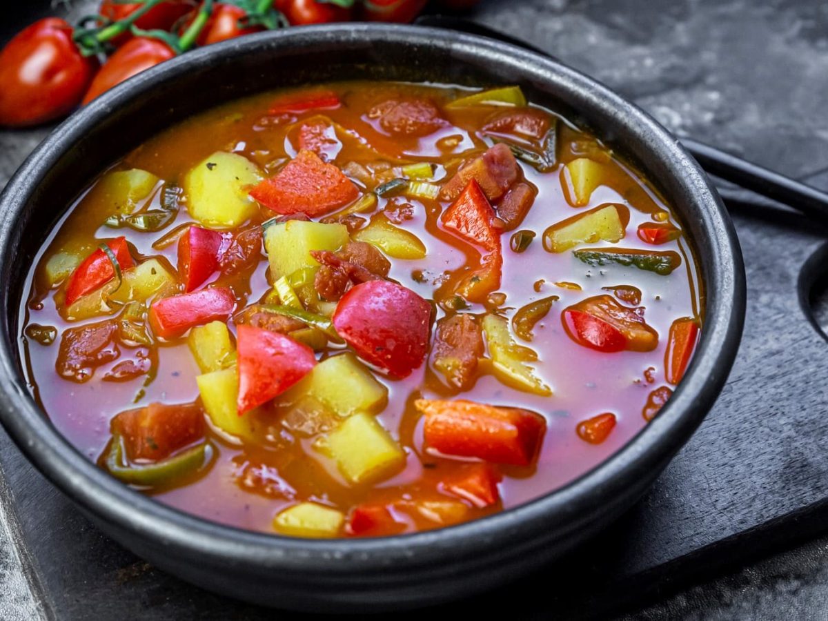 Eine schwarze Schale mitd er Gulaschsuppe auf einem Steinuntergrund und einem schwarzen Schneidebrett. Daneben ein kleiner Strauch mit Kirschtomaten.