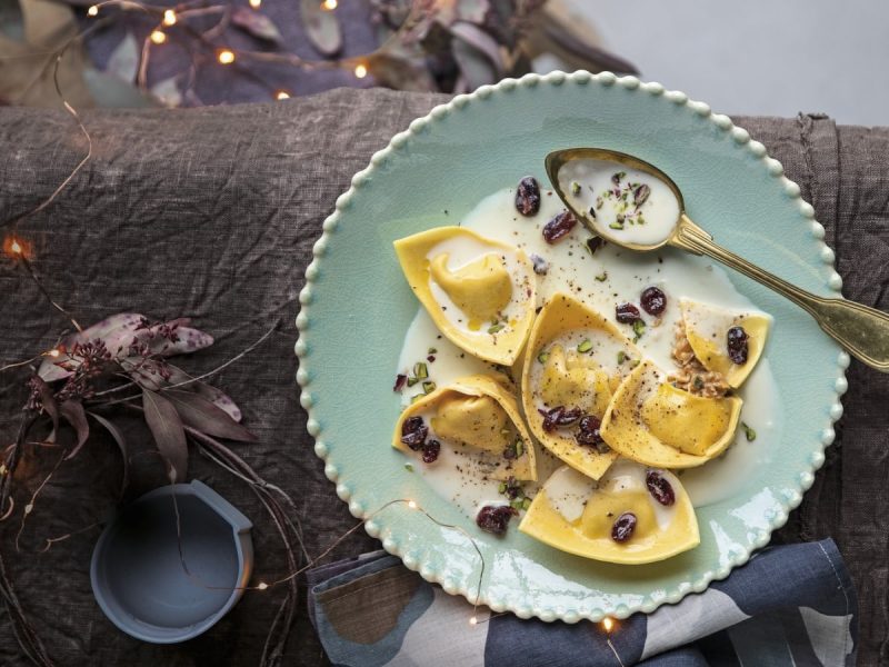 Draufsicht: Dunkler, hölzerner Untergrund mit dekorativer Lichterkette und Blumen. Außerdem ein türkisfarbener Teller mit sechs Ravioli in einer cremig hellen Sauce. Garniert mit getrockneten Cranberries und Pistazien.