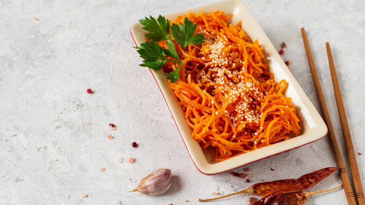 Ein weißer, rechteckiger Teller mit dem Kimchi aus Karotten auf grauem Untergrund. Daneben ein paar getrpcknete Chilis und etwas Knoblauch.