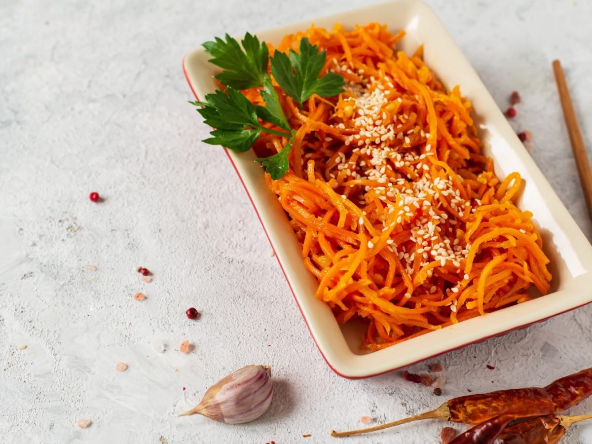 Ein weißer, rechteckiger Teller mit dem Kimchi aus Karotten auf grauem Untergrund. Daneben ein paar getrpcknete Chilis und etwas Knoblauch.