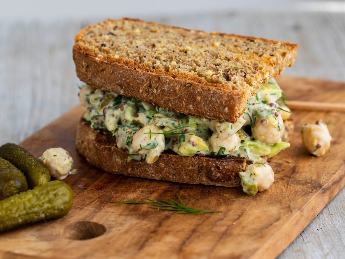 Ein Kichererbsen-Sandwich auf einem Holzbrett mit drei Mini-Gewürzgurken und etwas Dill.