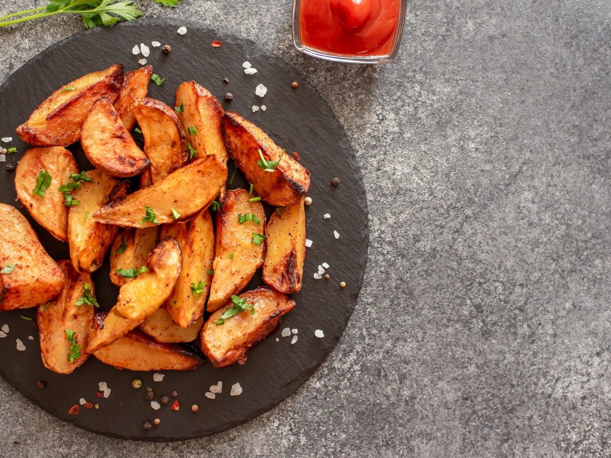 Kräuter-Knoblauch-Kartoffelecken auf einem Schieferteller, bestreut mit Petersilie und grobem Salz, in der Draufsicht.