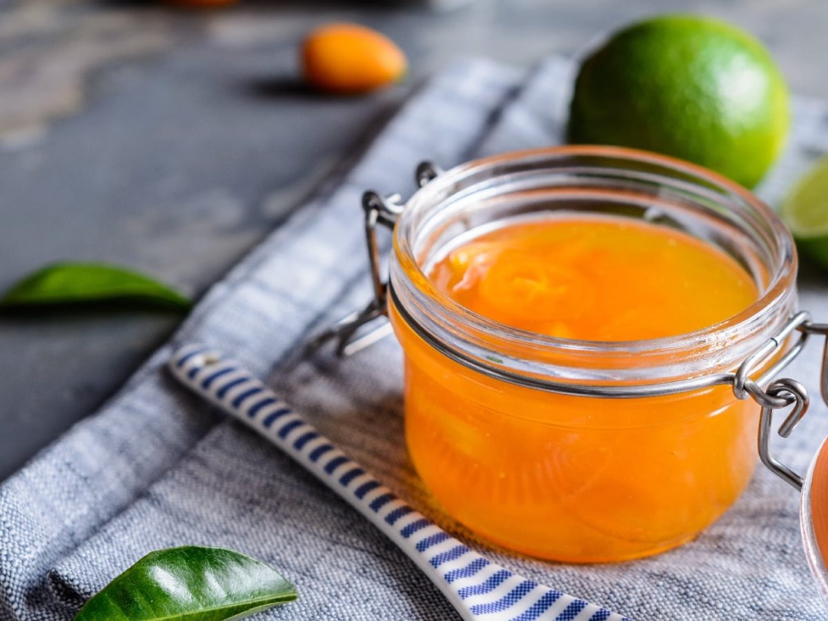 Ein Glas mit der Kumquat-Marmelade auf einem grauen Untergrund. Daneben eine Limette und Kaffirblätter.