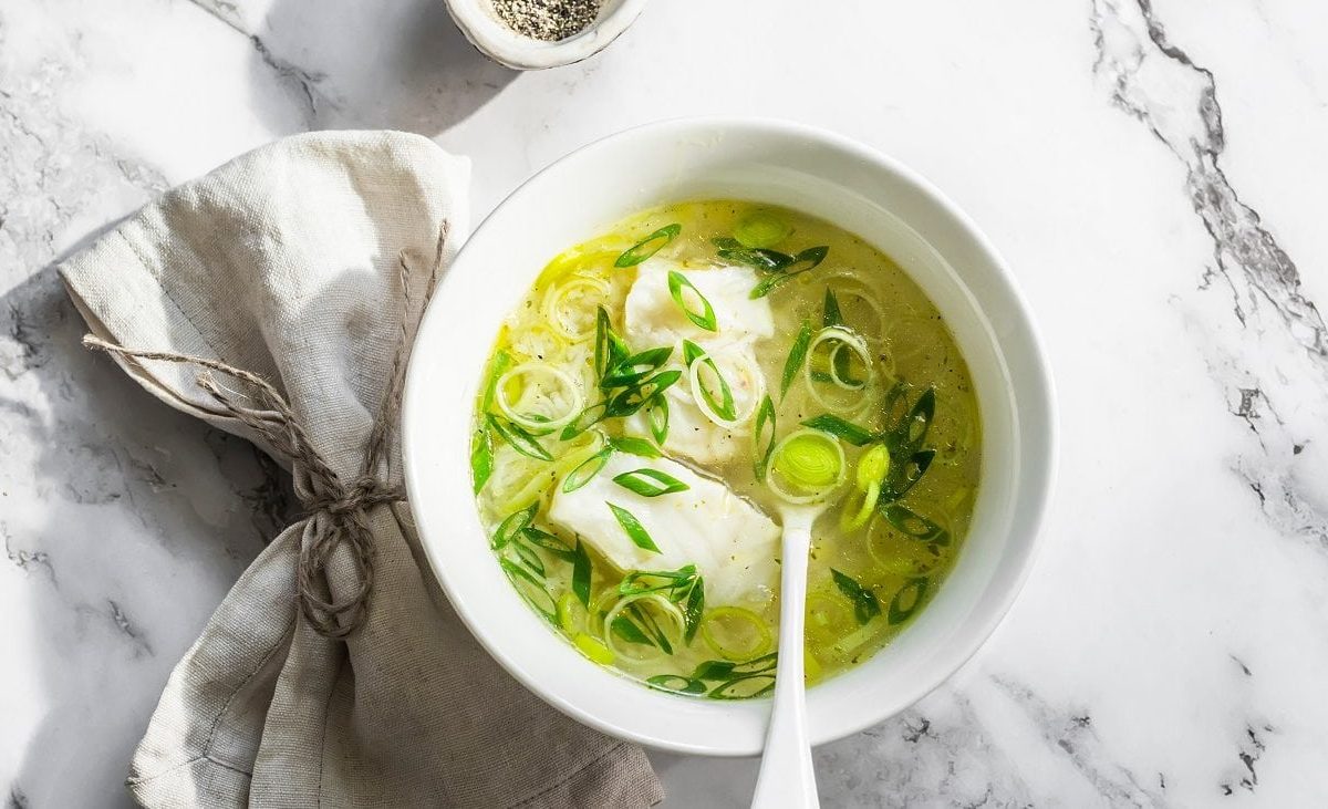 Draugsicht: eine helle Schale mit einer Leichte Lauch-Kabeljau-Suppe steht auf einen Untergrund aus hellem Marmor. In der Suppe ist ein Suppenlöffel.