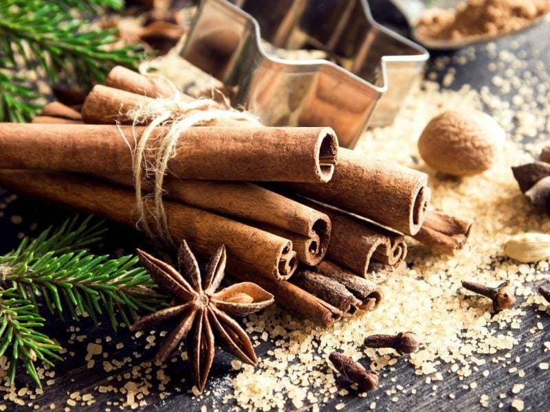 Lebkuchen-Gewürz., in seiner Rohform dargestellt durch Zimtstangen, Sternanis, Muskatnuss und braunen Zucker auf einem steinernen Tisch mit Tannennadeln.