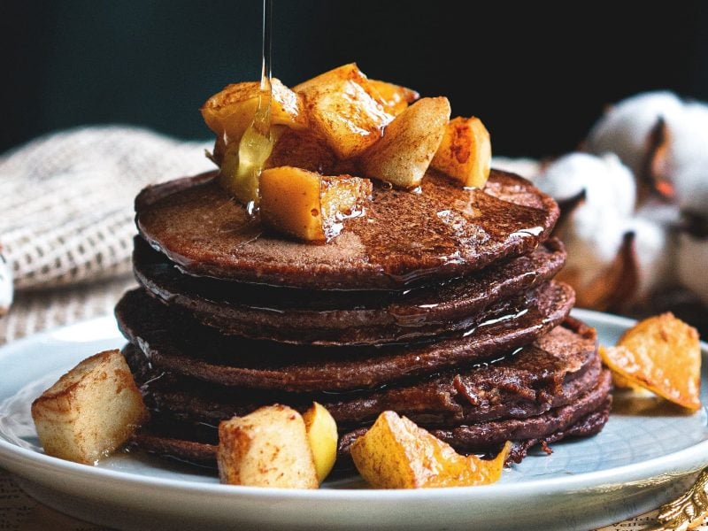 Ein Stapel Lebkuchen-Pancakes mit gebackenen Apfelstückchen und Sirup auf einem Teller.