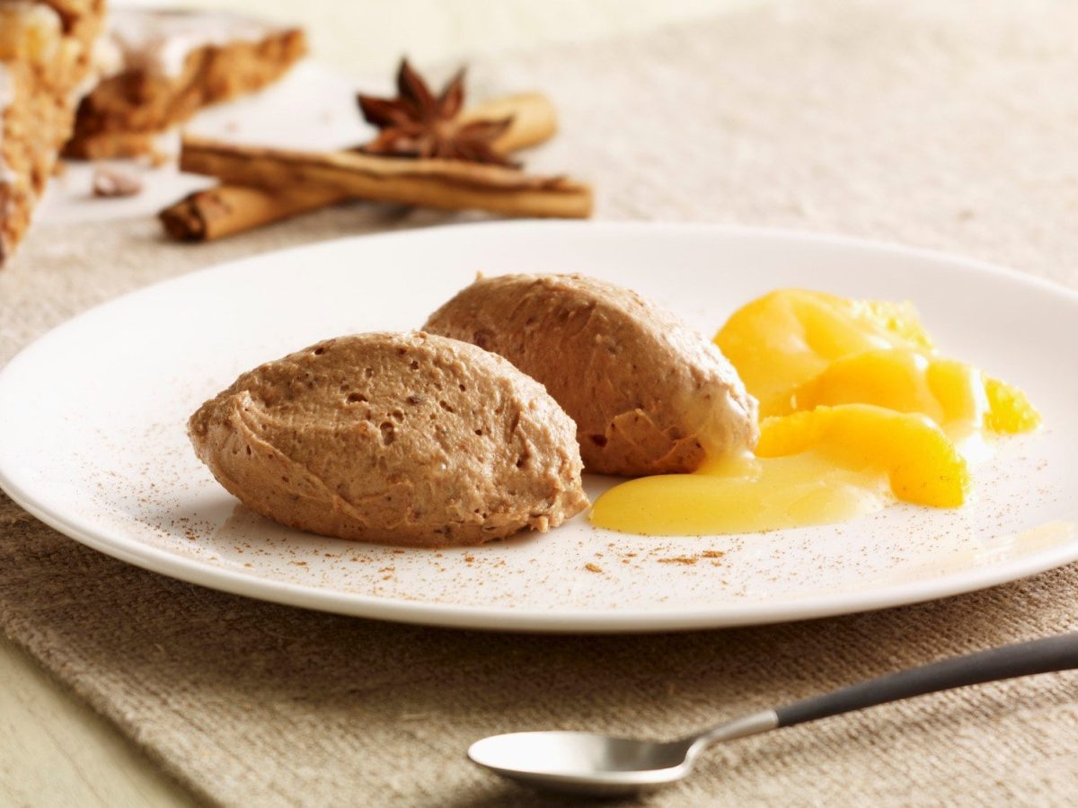 Ein weißer teller auf beigem tisch mit der Lebkuchenmousse und den Orangen. Links daneben Lebkuchen, Zimt und Sternanis