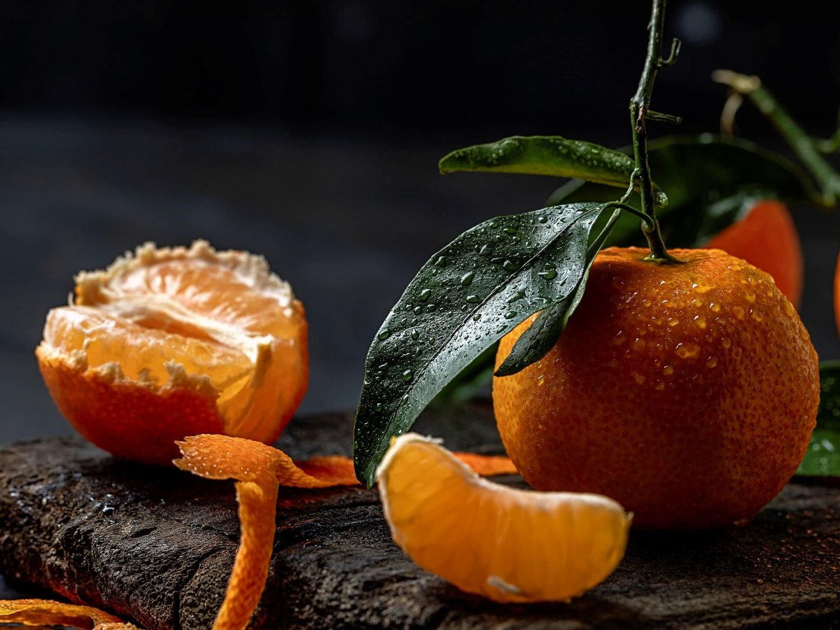 Mandarinen aufbewahren mit Mandarinen mit Grün auf Holzplatte auf dunklem Untergrund. Im Bild Mandarinenschale und einzelne Stücke. Frontalansicht.