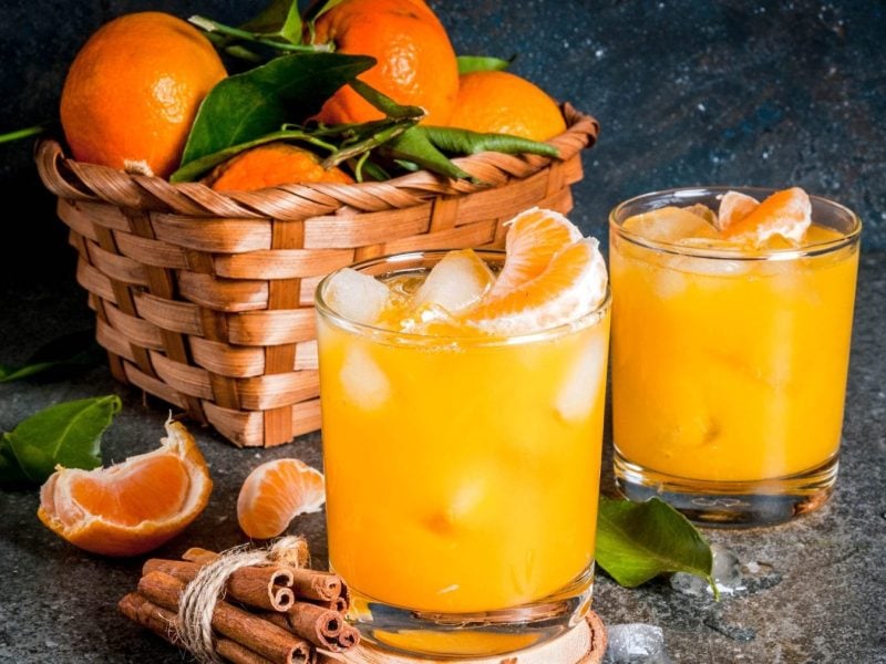 Mandarinen Cocktail mit Vodka, Zimt und Anis in zwei Gläsern mit Eis. Dahinter eine Schale mit Mandarinen. Im Bild Mandarinenstücke und Zimtstangen. Frontalansicht.