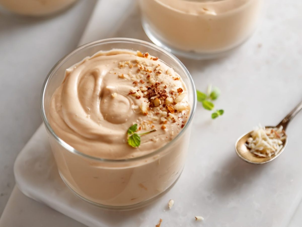 Zwei Gläser mit der hellbraunen Mandel-Mousse auf einem weißen Tisch. mit einem langen, kleinen silbernen Löffel.