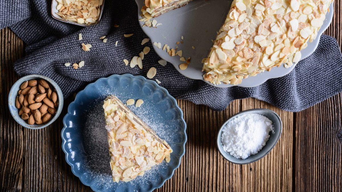 Ein Stück Mandeltorte auf einem kleinen blauem Teller. Daneben der Rest des Kuchens auf einem dunklen Holztisch mit blau-grauen Tischtüchern.