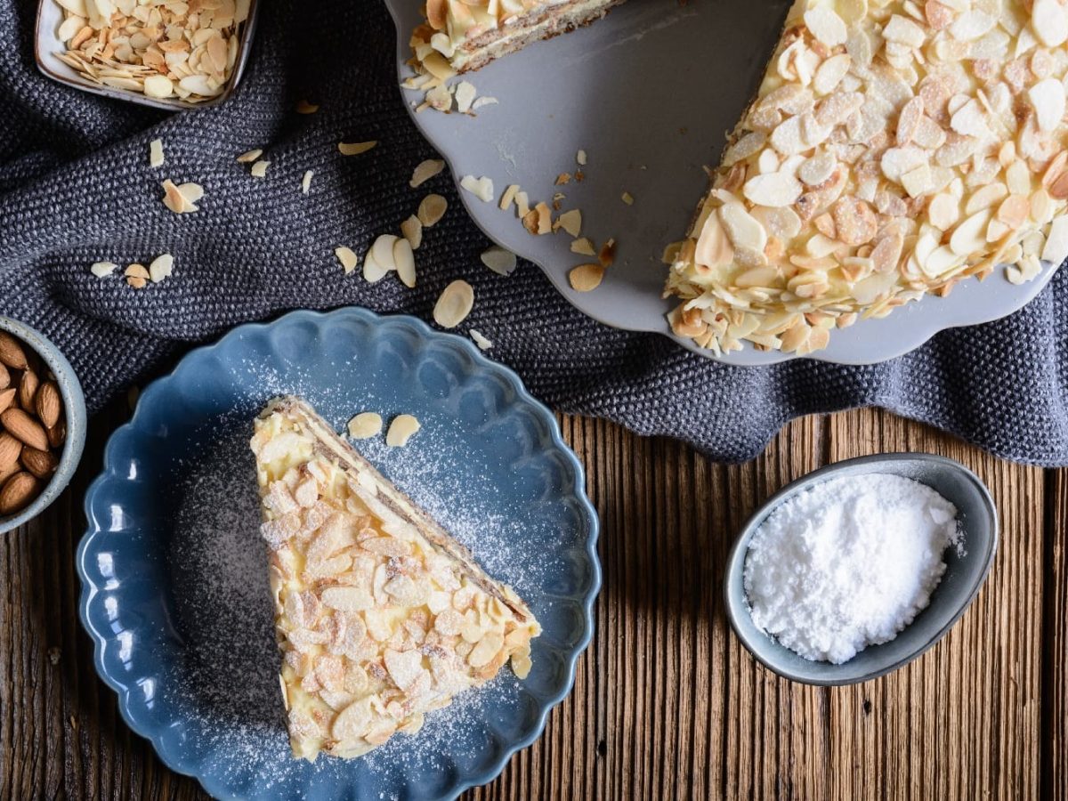 Ein Stück Mandeltorte auf einem kleinen blauem Teller. Daneben der Rest des Kuchens auf einem dunklen Holztisch mit blau-grauen Tischtüchern.