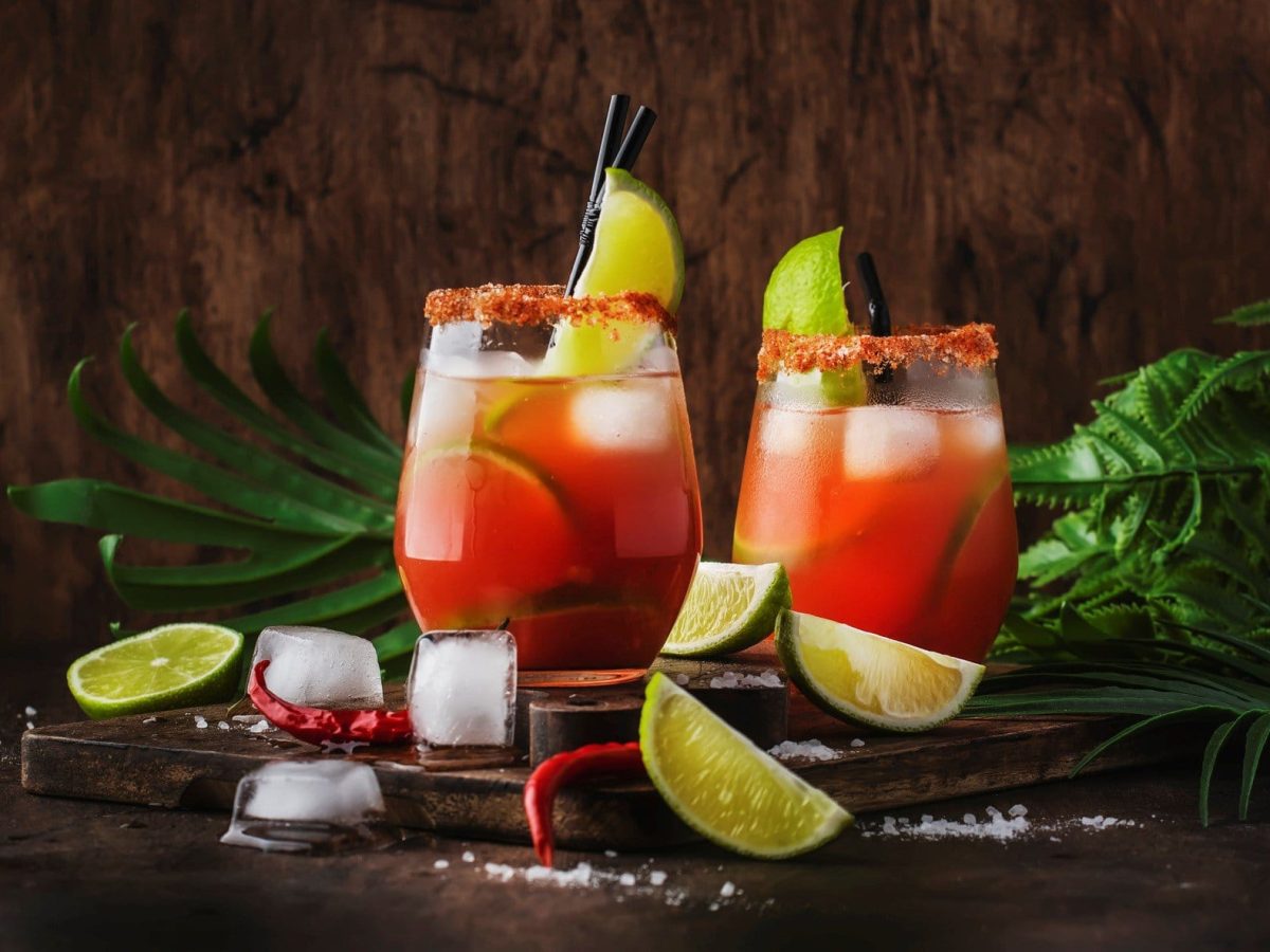 Michelada mexikanischer Biercocktail mit Lager in zwei Gläsern auf dunklem Holz vor dunklem Hintergrund. Im Bild Pflanzen, Limettenspalten, Salz, Chilis und Eiswürfel. Frontalansicht.