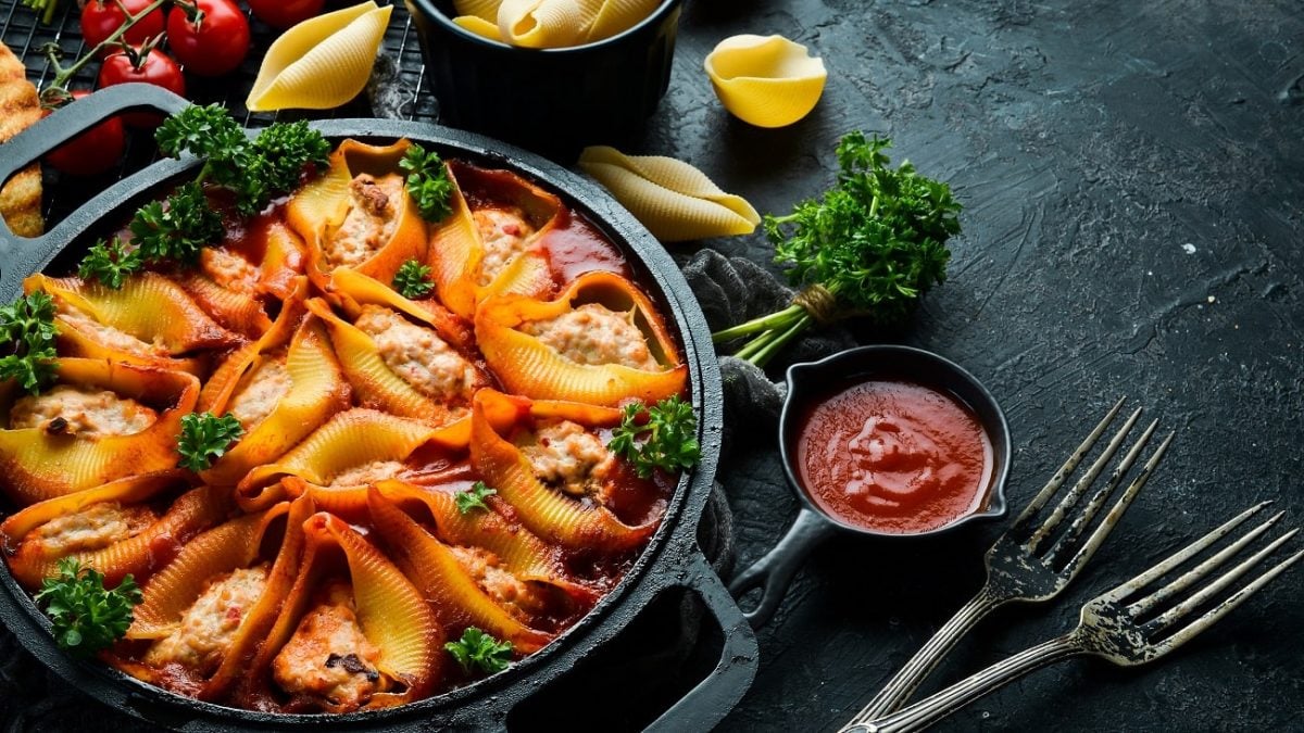 Draufsicht: Auf einem dunklen Hintergrund steht eine Pfanne mit Muschelnudeln mit Petersilien-Hack-Füllung. Drum herum liegen zwei Gabeln, eine Tomatensoße, Petersilie, Tomaten und trockene Muschelnudeln.