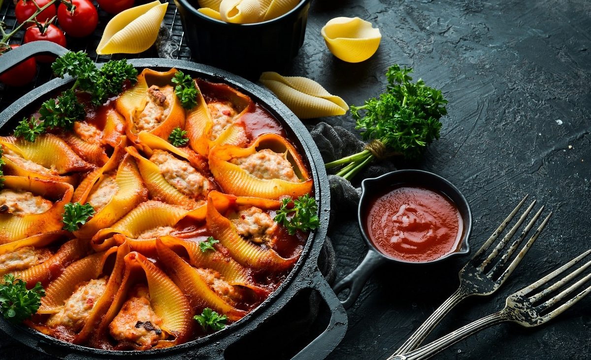 Draufsicht: Auf einem dunklen Hintergrund steht eine Pfanne mit Muschelnudeln mit Petersilien-Hack-Füllung. Drum herum liegen zwei Gabeln, eine Tomatensoße, Petersilie, Tomaten und trockene Muschelnudeln.