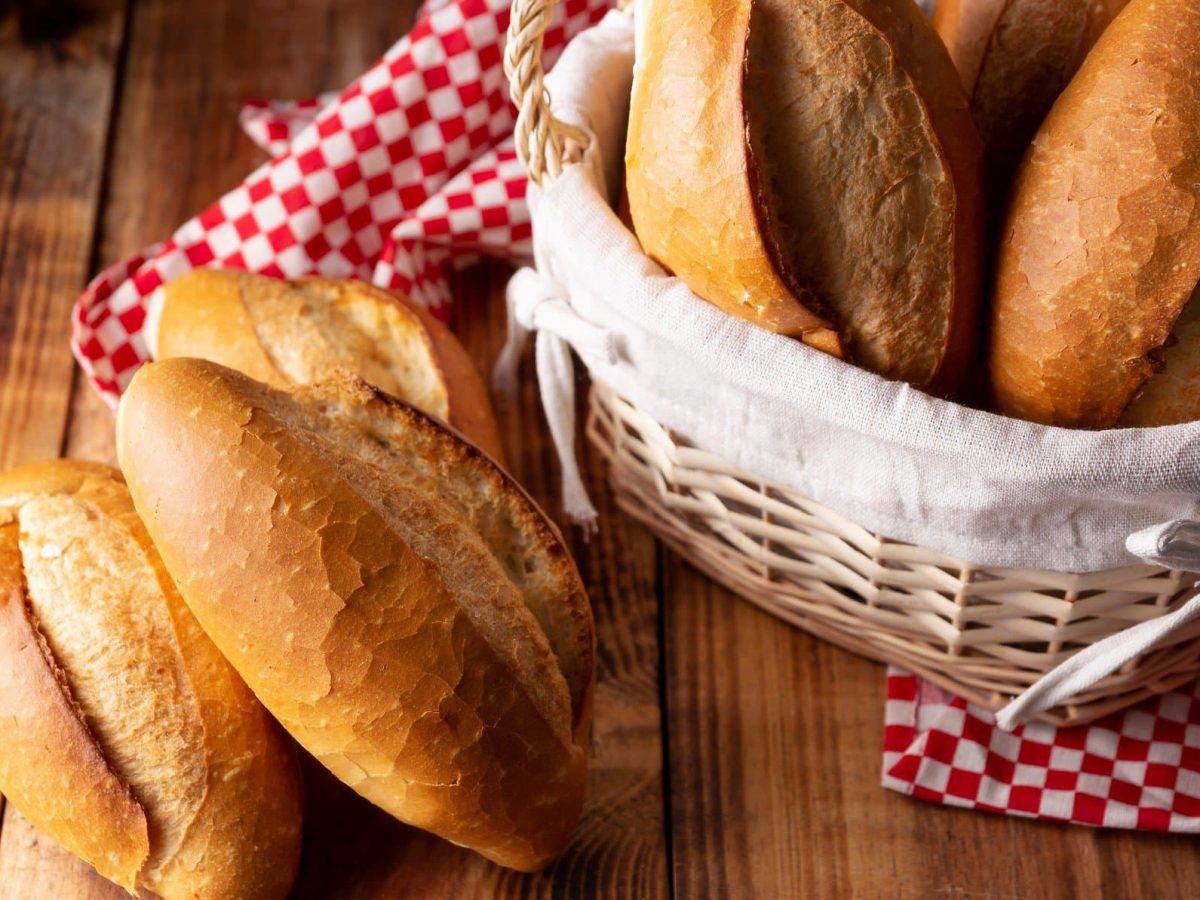 Original mexikanische Bolillos Brötchen wie Baguette in einem Korb mit rot-weißem Tuch auf Holztisch. Daneben weitere Bolillos. Draufsicht.