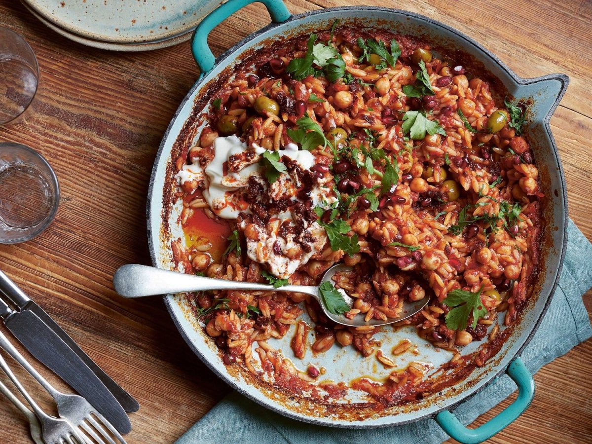 Eine Auflaufform Orzo mit Kichererbsen und Oliven, darüber frische Kräuter, in der Draufsicht.