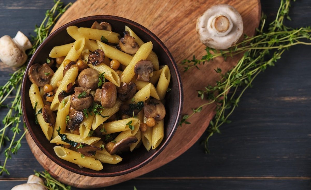 Draufsicht: Auf einem Holzbrettchen steht eine schwarze Schüssel mit Pasta mit Thymian-Champignons. neben dem Brettchen liegen rohe Champignons und mehrere Zweige frischen Thymian.