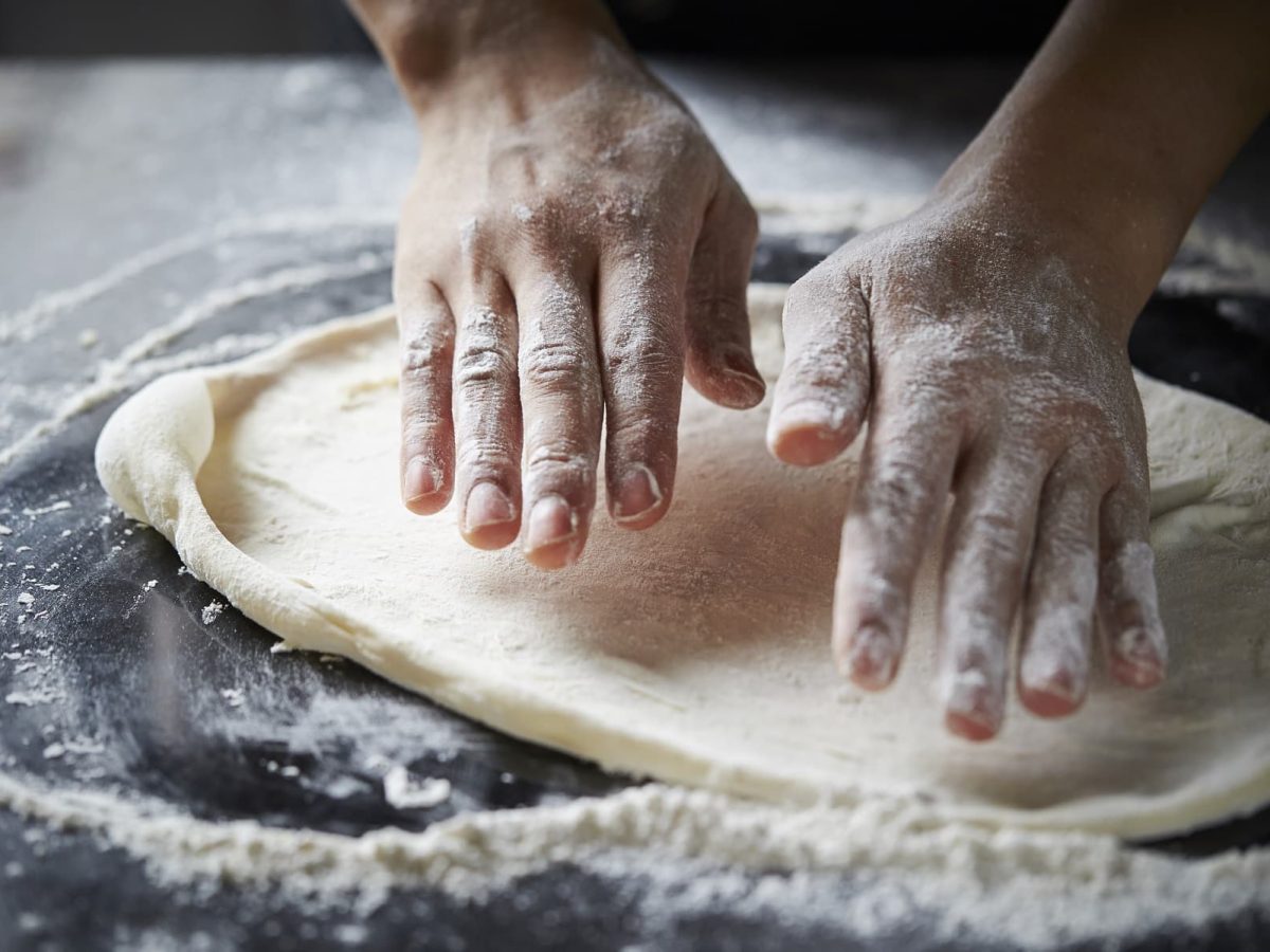 Frontalsicht: Auf einer dunklen Holzplatte ist mehl verstreut. Zwei Hände kneten einen Pizzateig, der fast rund ist.