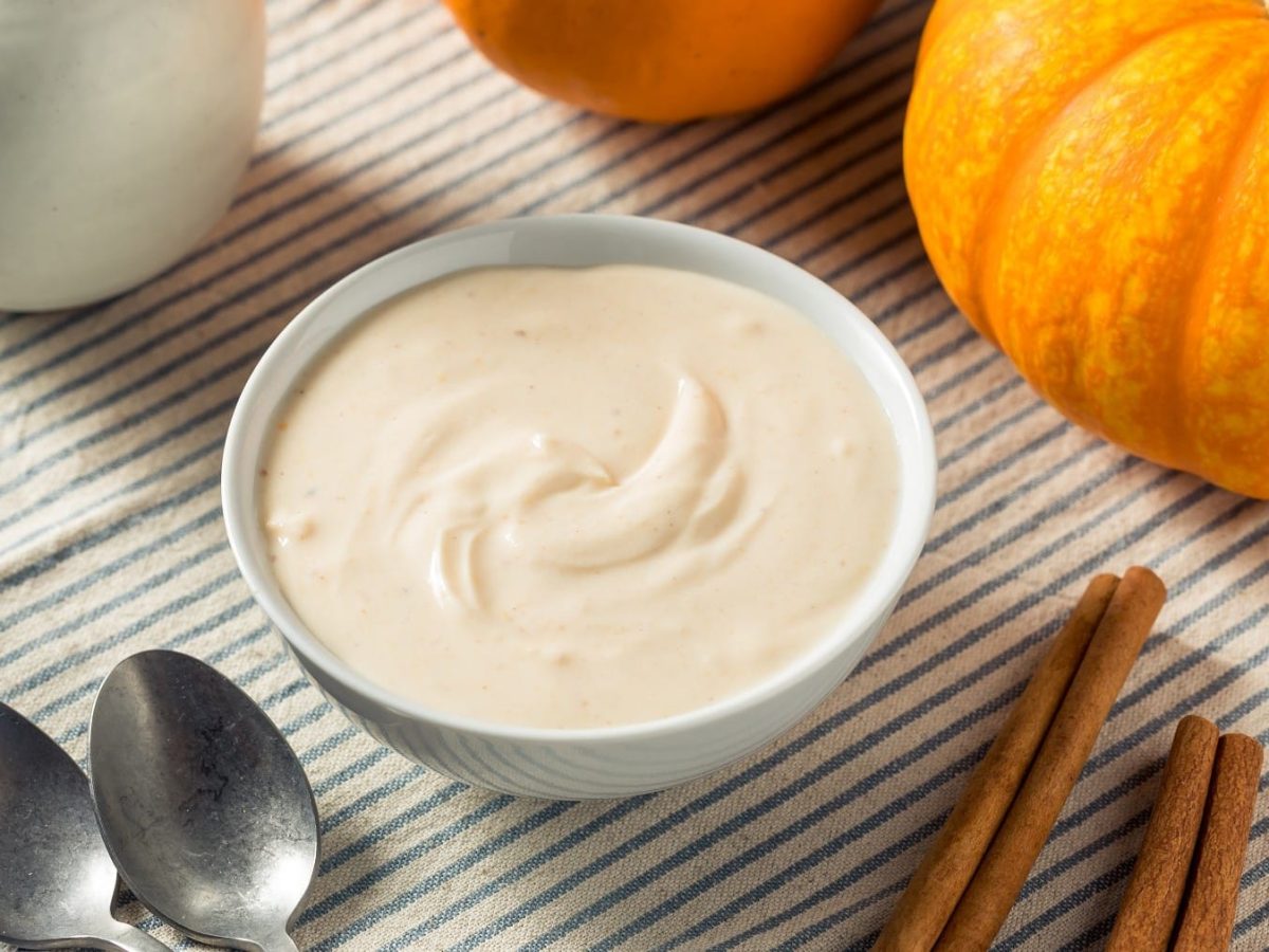 Pumpkin Spice Joghurt, serviert in einer Schale auf einem einfach gedeckten Küchentisch mitsamt ein paar Stangen Zimt und einem Hokkaidokürbis.